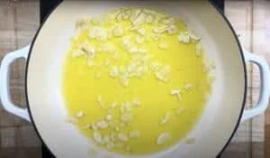 Sliced garlic sizzling in olive oil inside a white Dutch oven on a wooden counter.